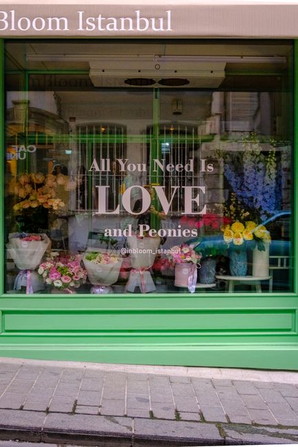 The image shows a flower shop window display featuring a variety of fresh floral arrangements with vibrant colours, including pink, yellow, purple, and white flowers. The bouquets are neatly wrapped i