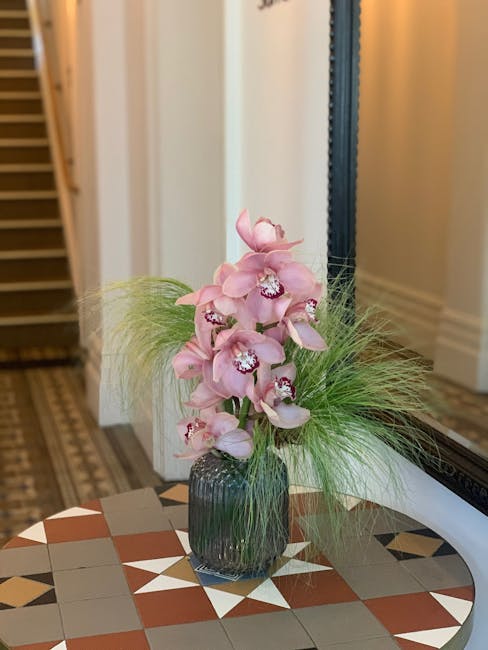 A tall floral arrangement featuring soft pink orchids with multiple blooms on each stem, set in a transparent, ribbed glass vase placed on a decorative table with geometric tile patterns. The orchids 