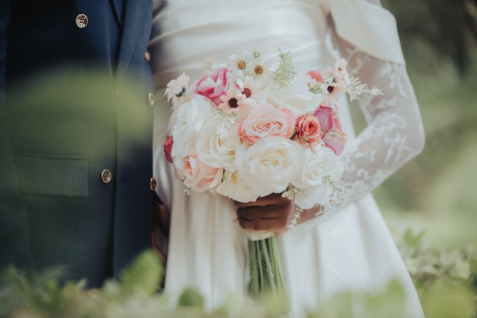 Close-up of a bridal bouquet featuring a mix of white and blush pink roses, accented with small white daisies and green foliage, held by a bride in a white lace wedding dress. The bouquet is arranged 