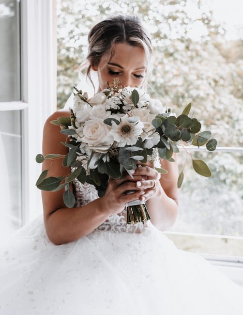 A bride holding a large, elegant bouquet of fresh flowers, featuring white roses, white anemones with dark centers, and various greenery including eucalyptus leaves. She is seated indoors near a windo
