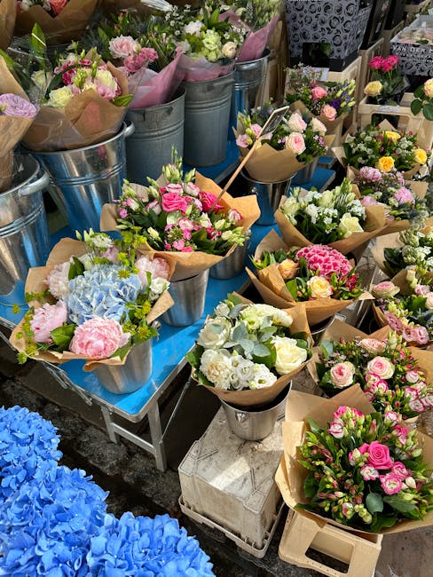A display of freshly arranged floral bouquets and potted plants at a flower market, featuring soft pink roses, white hydrangeas, pale green foliage, and blush peonies wrapped in brown craft paper, arr