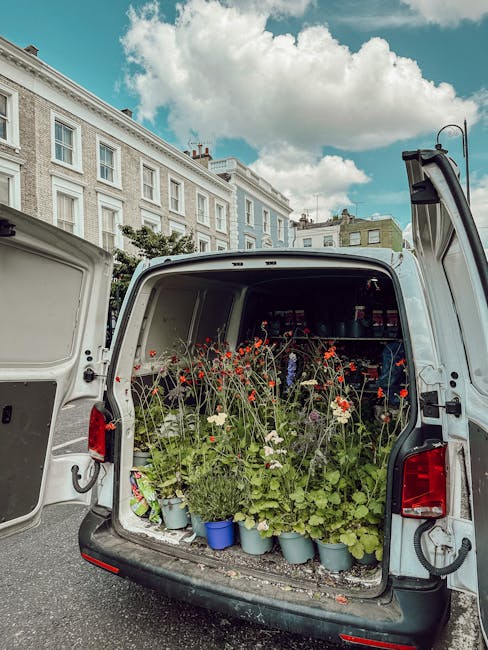 The open back of a white delivery van filled with potted plants and flowering garden flowers, including red poppies, white cosmos, and green foliage, arranged on shelves inside. The van is parked on a