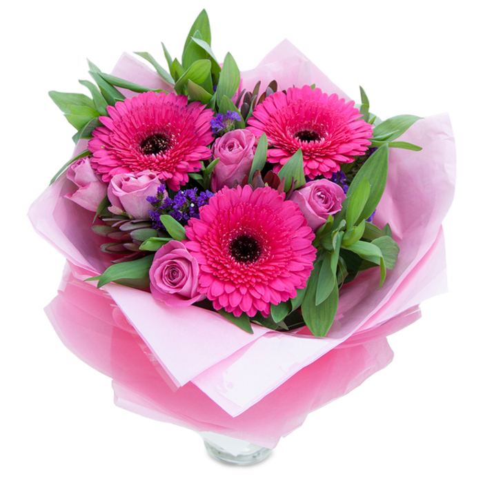 As a Bloomsbury florist I imagine this bouquet sitting beneath the soft light that filters in from Russell Square - three striking fuchsia-pink Gerbera daisies form the focal point, their radiating petals and dark centres lending a bold graphic contrast. Around them, roses in a muted lavender-pink or dusty-rose unfurl with delicate, velvety curves, each bud still holding the freshness of recent harvest. Among these larger blooms are pockets of tiny purple statice or limonium, adding an airy, almost wildflower texture that breaks up the mass of petals and introduces a cooler violet tone. A generous sweep of green foliage, suggesting salal and eucalyptus, gives the arrangement depth and a crisp, botanical scent that balances the rose perfume. The bouquet is wrapped with mindful craftsmanship: an inner layer of pale pink matte tissue paper cradles the stems while an outer veil of translucent, slightly crinkled fuchsia-pink cellophane provides a luminous sheen and keeps the colour story cohesive. The wrapping's folds and the exposed rim of a clear glass vase hint at a piece designed for immediate display - ideal for a mantel, bedside table or as a centrepiece at a small Bloomsbury gathering near the British Museum or Brunswick Centre. Light plays across petal edges and cellophane, and the combined textures - velvety petals, crisp foliage, papery statice - invite touch. Available for local delivery from Flowers Bloomsbury, this bouquet feels celebratory yet intimate, suited to birthdays, Mother's Day, anniversaries, or simply to make someone feel cherished.